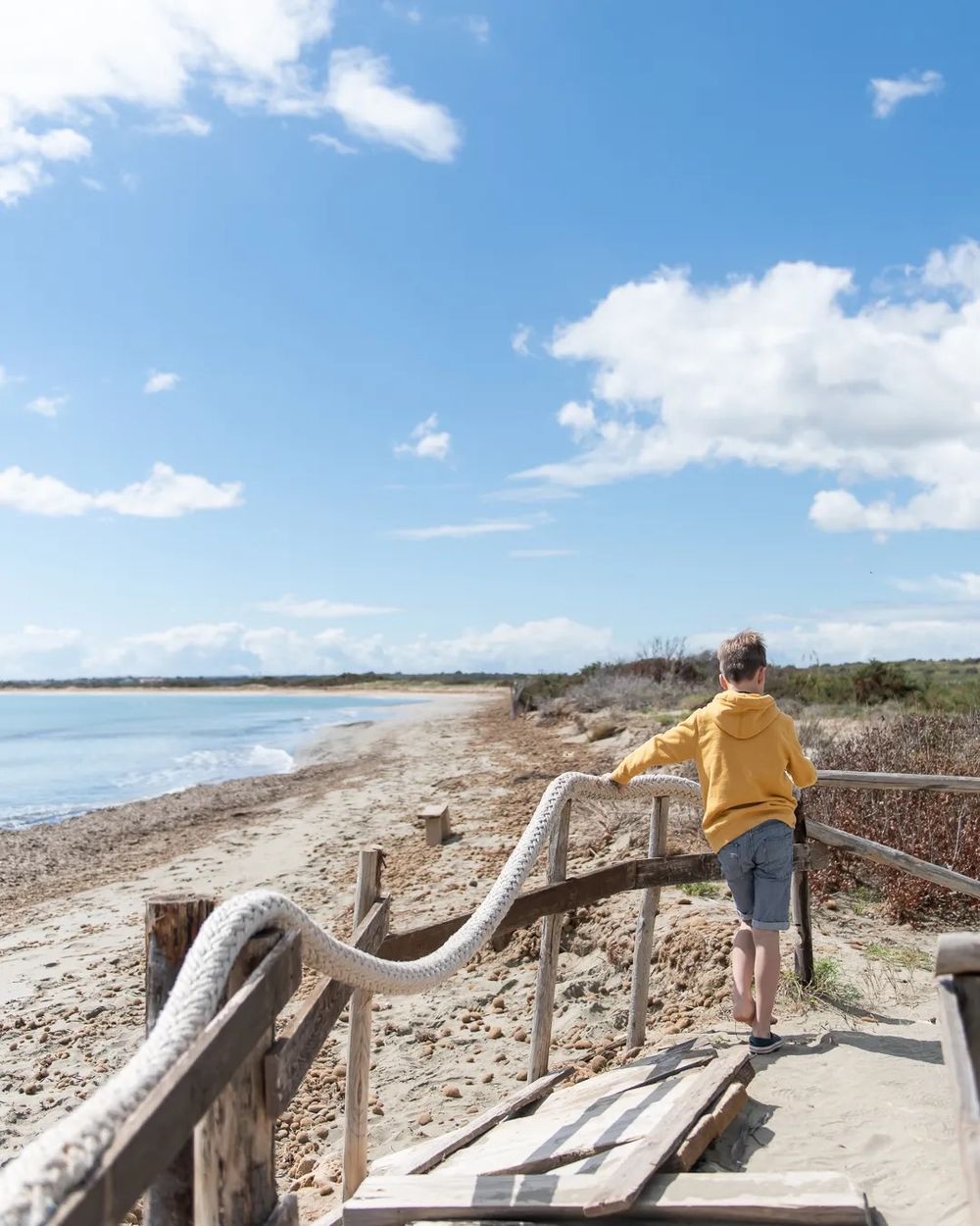 A child standing on a wooden coastal boardwalk overlooking the beach under a bright blue sky.