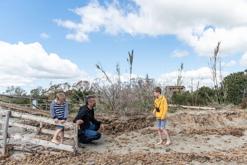 Two children and an adult work together near sand dunes under a bright cloudy sky.
