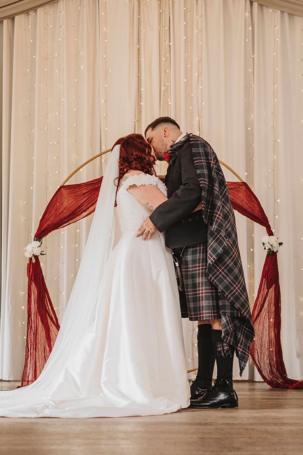 Wedding couple in Scottish attire sharing an intimate moment against a backdrop of fairy lights and curtains.