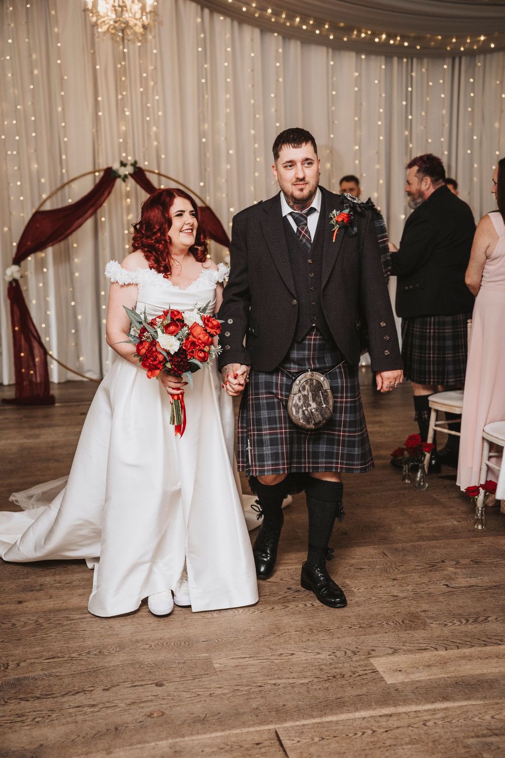 Wedding party walking together in a reception hall decorated with fairy lights and elegant chandeliers.