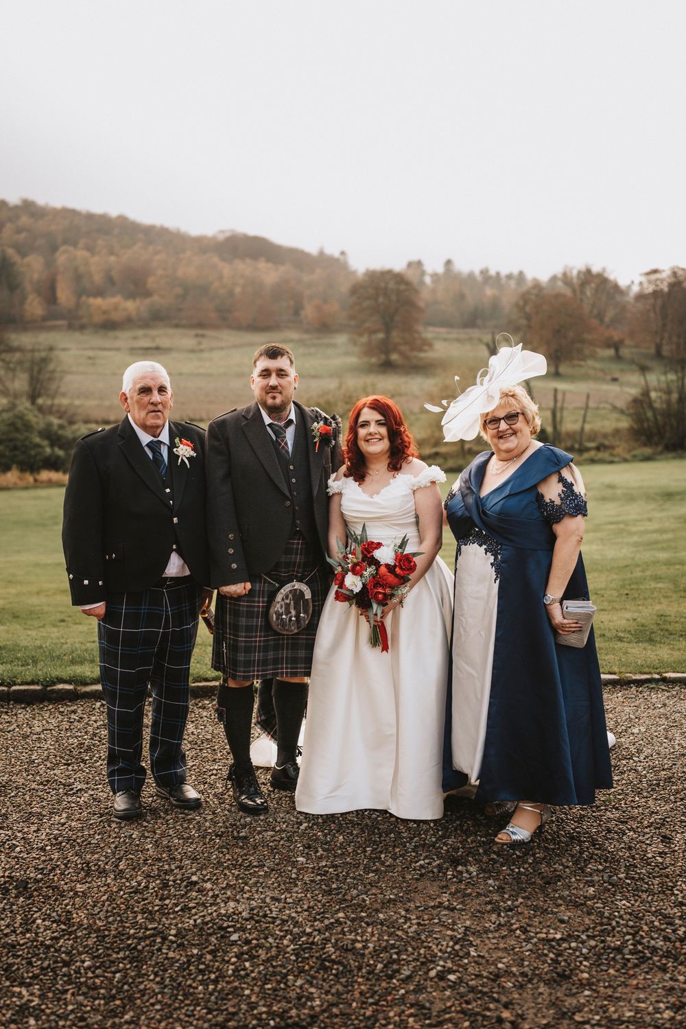 Wedding party in traditional Scottish kilts and formal attire pose together on gravel path with rolling hills behind.