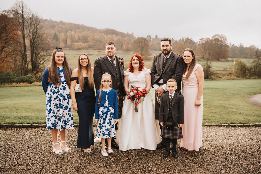 Wedding party group photo taken outdoors with scenic autumn landscape in background.