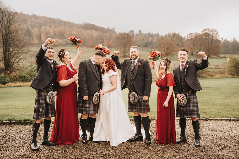 Wedding party in burgundy dresses and kilts posing for photos in an autumn outdoor setting.