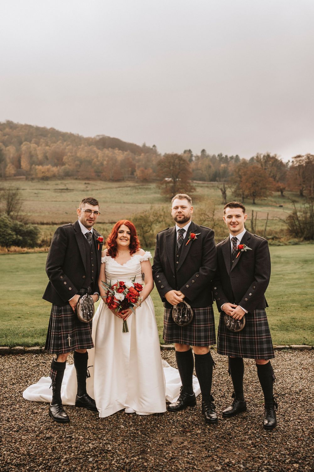 Wedding party in traditional Scottish kilts and formal attire posing outdoors against a rural landscape.