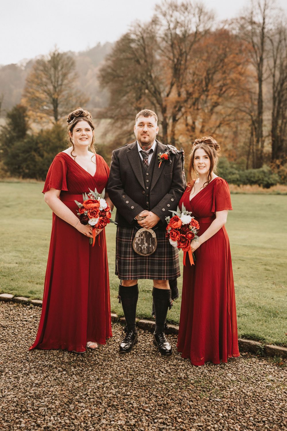 Wedding party poses outdoors in autumn with bridesmaids in red dresses and groom in traditional Scottish kilt.