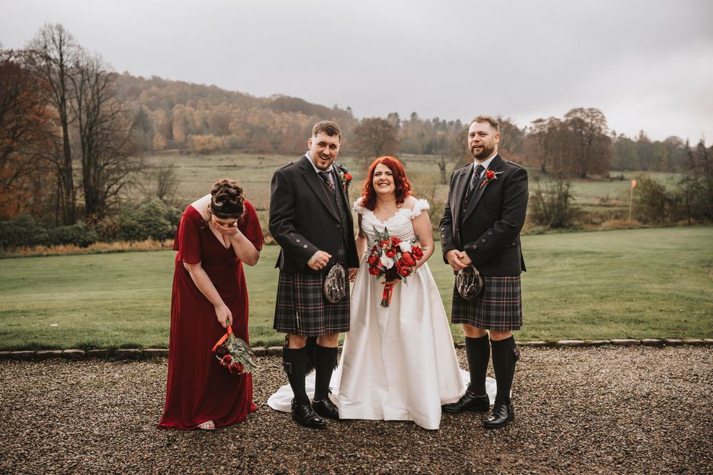 Wedding group photo on a gravel path with Scottish countryside and autumn trees in background.