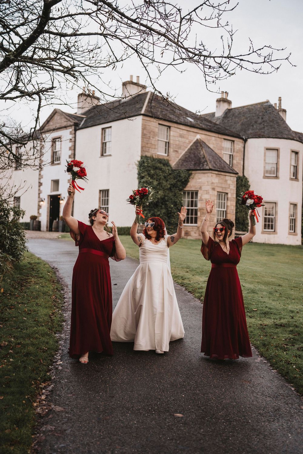 Bridal party in burgundy dresses celebrating outside a traditional Scottish stone building.