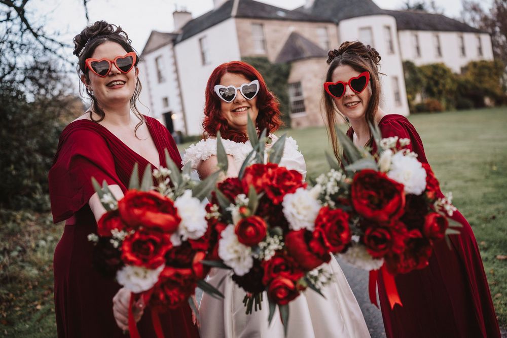 Wedding party holding vibrant red and white bouquets in front of a rustic building.