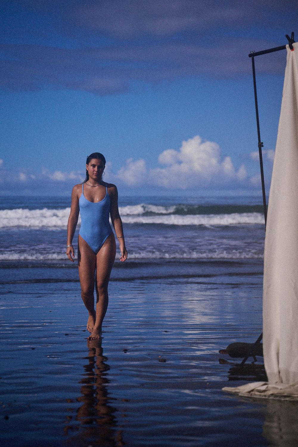 A women in a blue swimsuit walks on a reflective beach at low tide during a swimwear photoshoot by photographer Rachael Kershaw.
