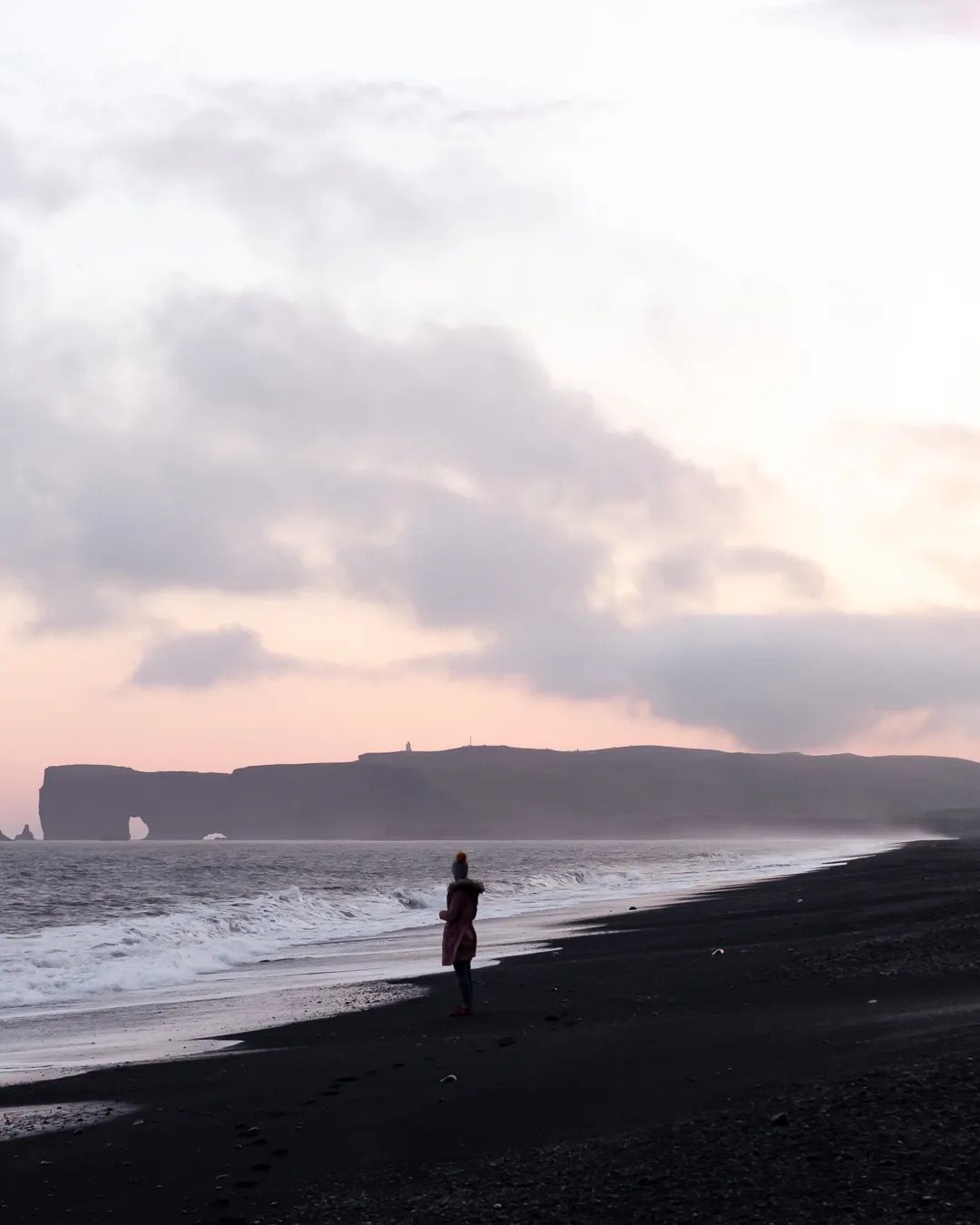 Person walking along a black sand beach under a pastel sunset sky.