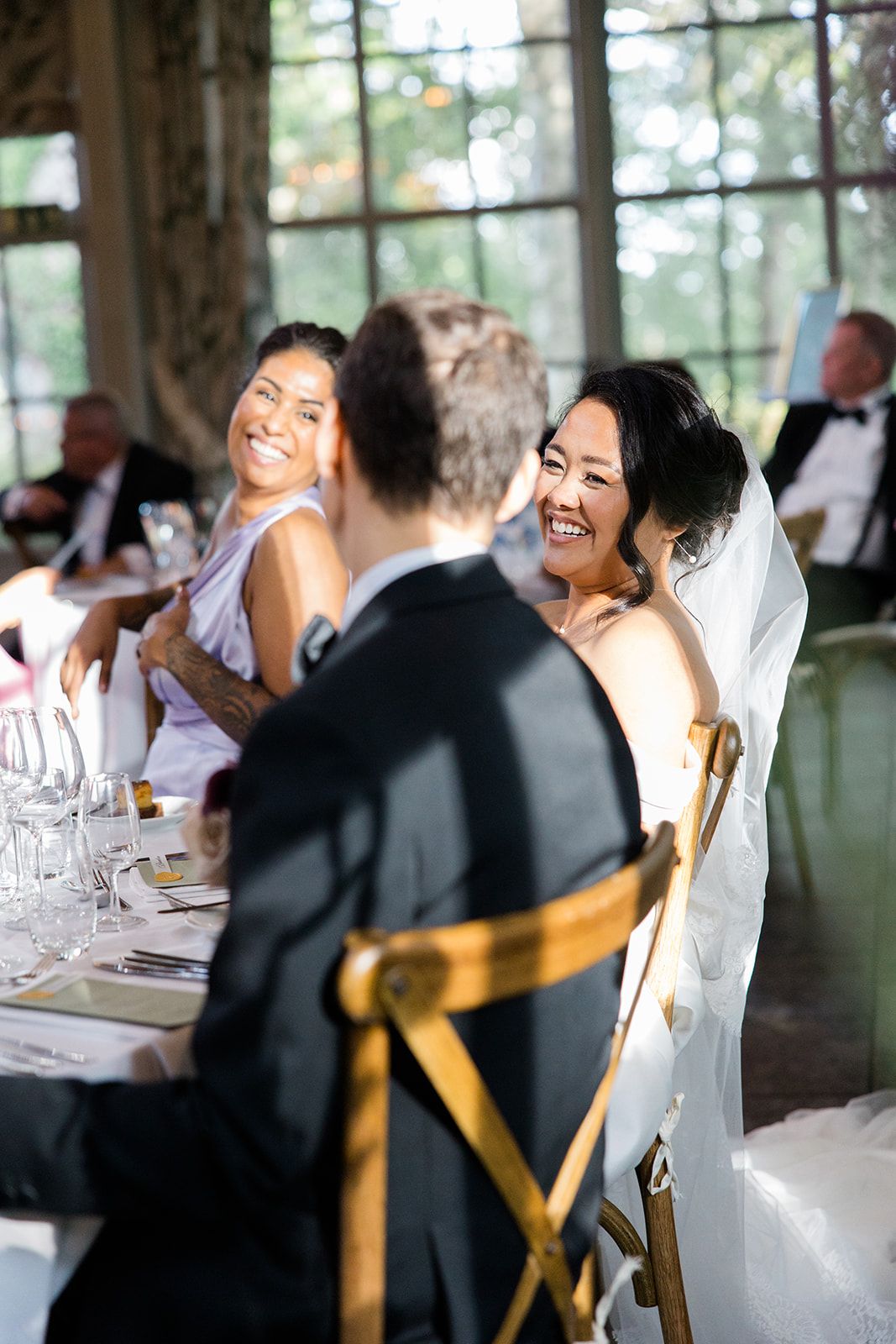 Wedding guests share joyful laughter during reception dinner at elegant indoor venue with natural lighting.