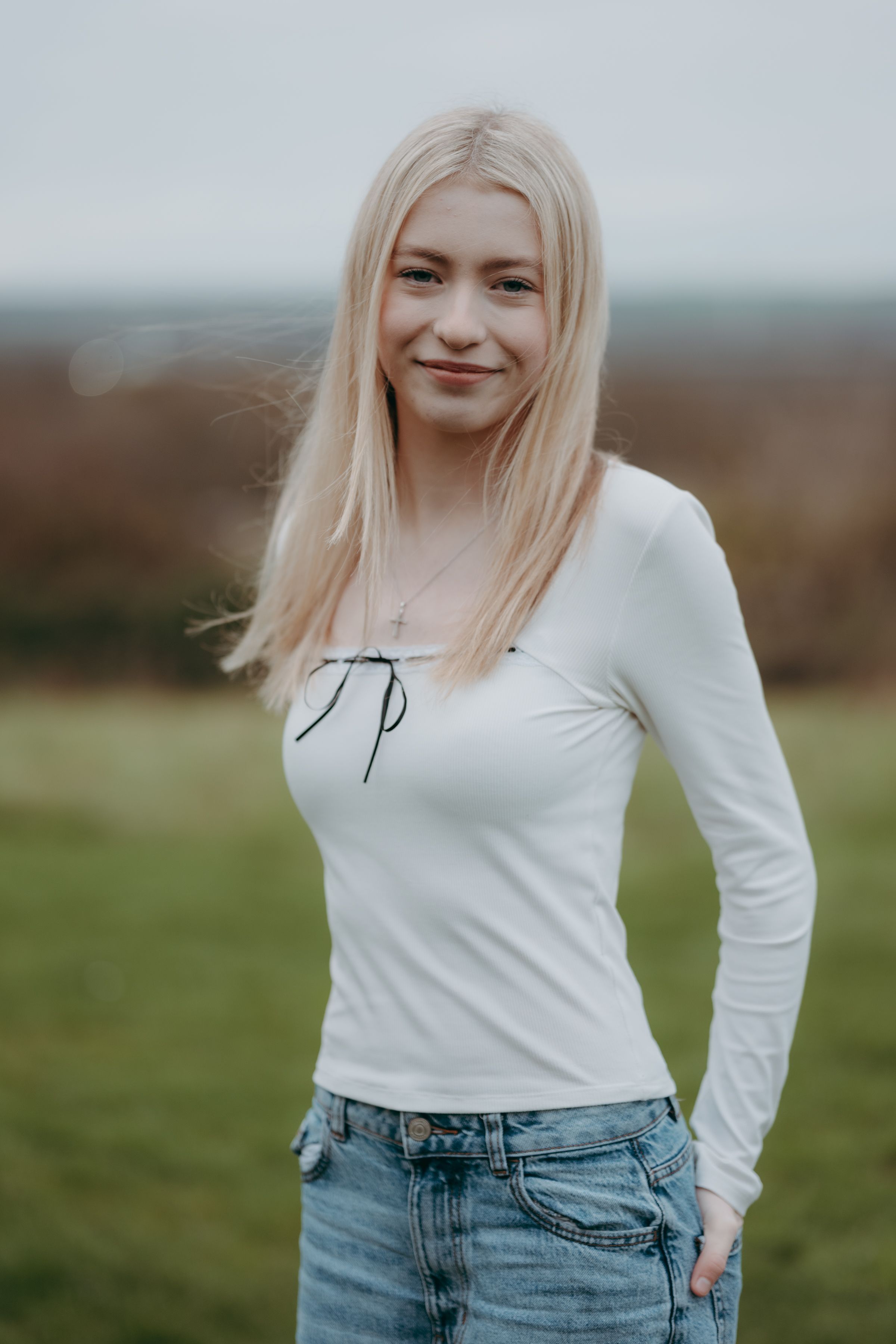 Person in a white long-sleeve top and blue jeans poses against a blurred outdoor background.