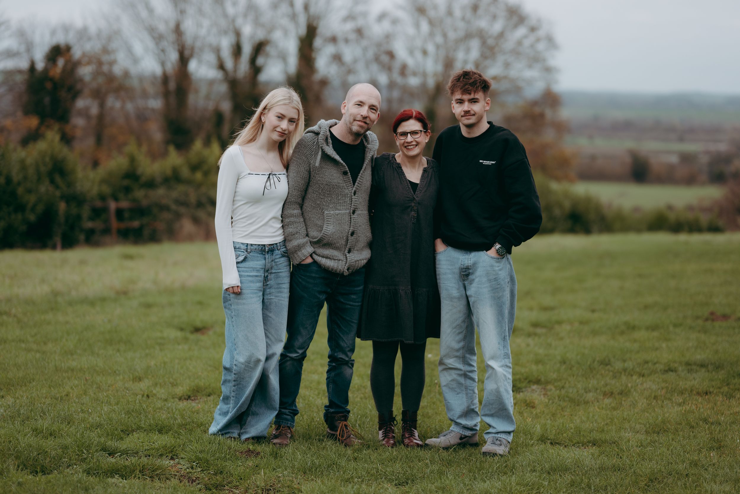A group of four people standing together on a grassy hill during autumn with trees in the background.