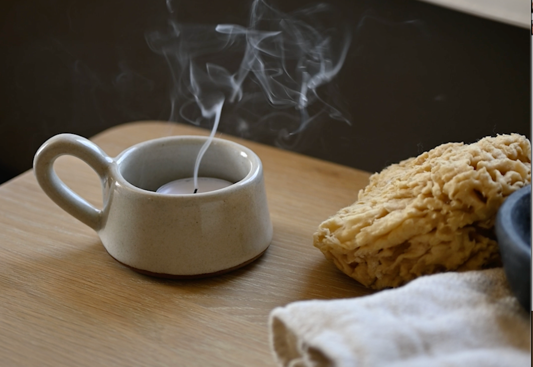 Smoke rises from the candle holder next to a natural sponge and washcloth in the beautiful bathroom setting