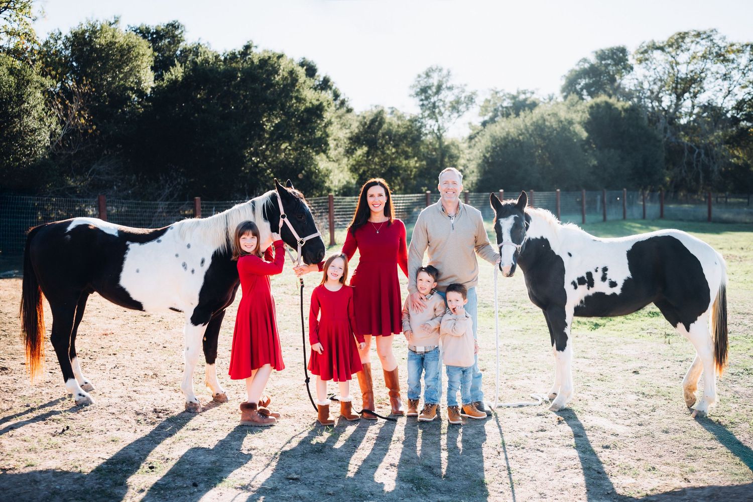 A family in red dresses and casual attire poses with horses together in a sunlit outdoor setting for a photo session.