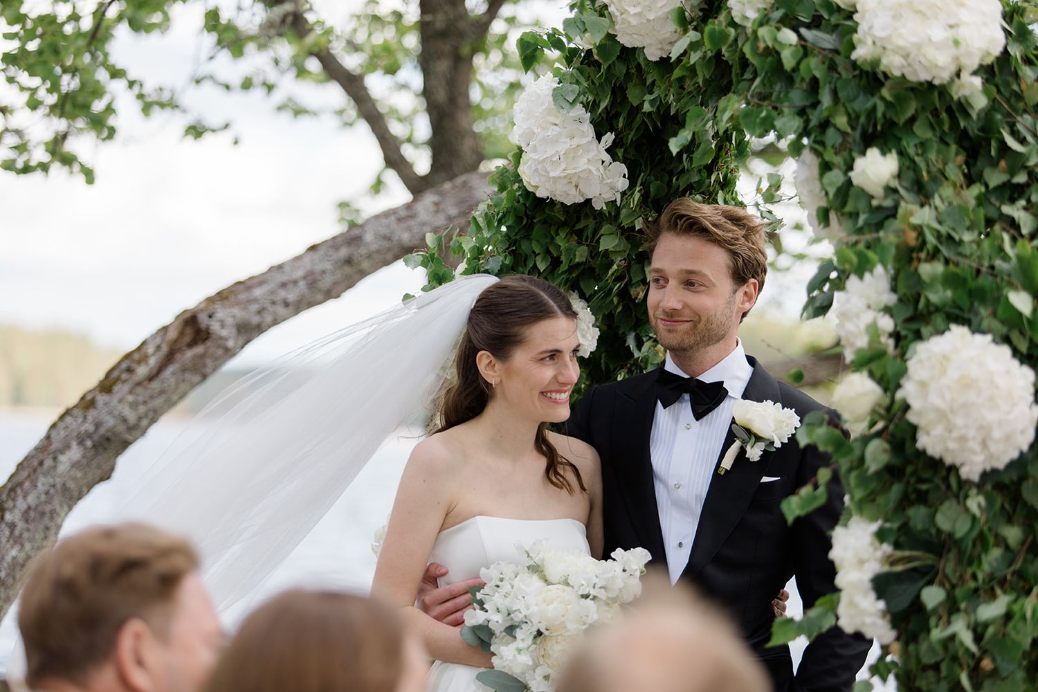 A wedding couple embraces under lush green trees and white floral arrangements during an elegant outdoor ceremony.