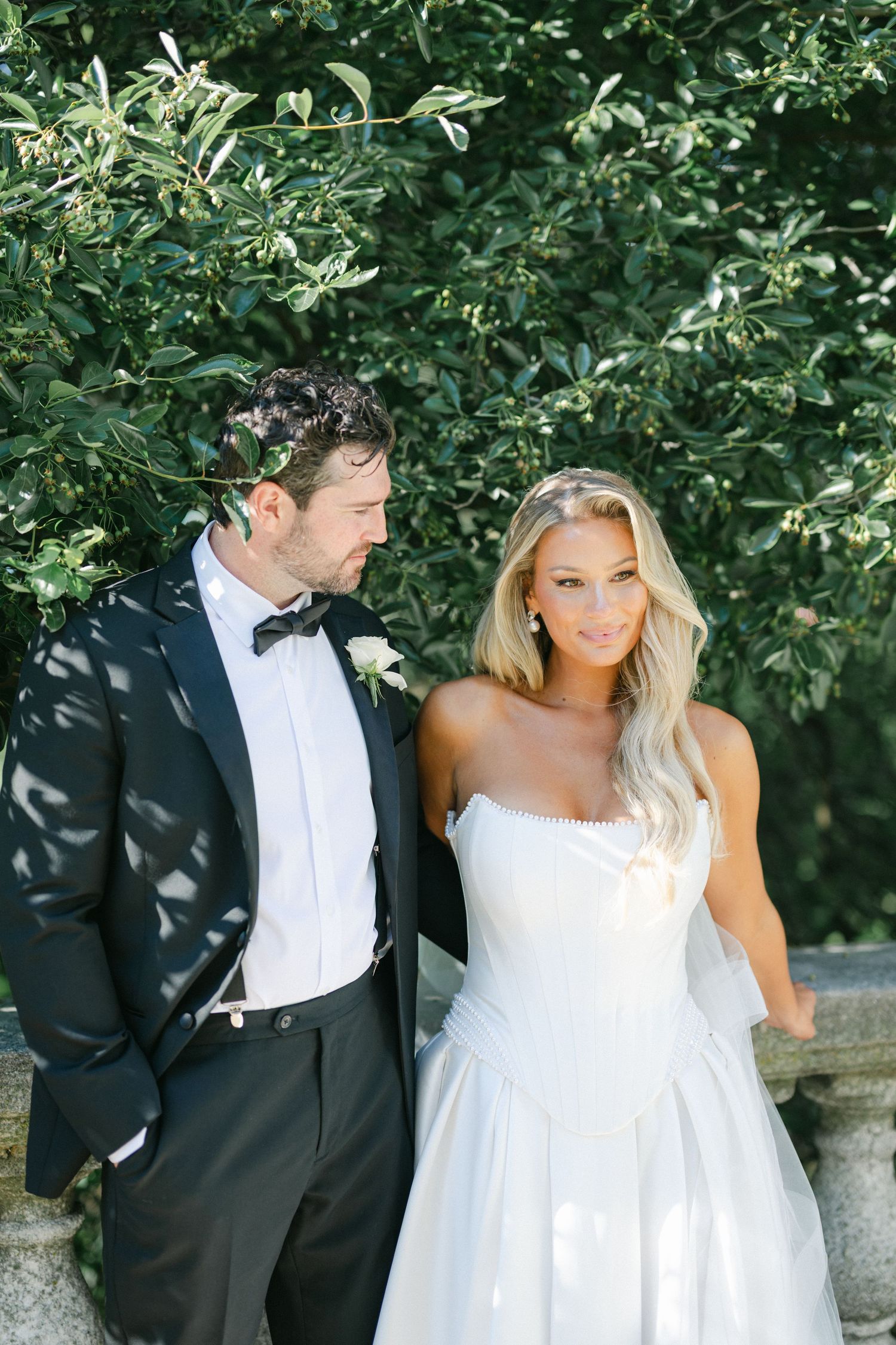 Bride in white strapless dress with lace trim holds bouquet of white roses in outdoor garden setting.