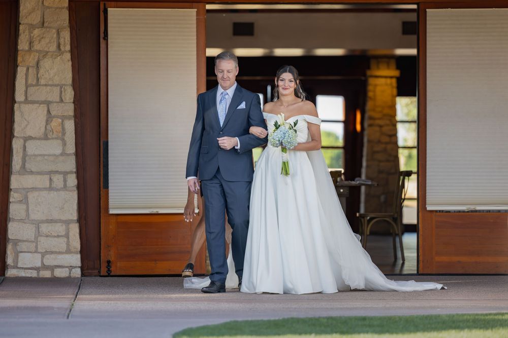 Father escorts bride in an elegant off-shoulder wedding gown through venue entrance doors.