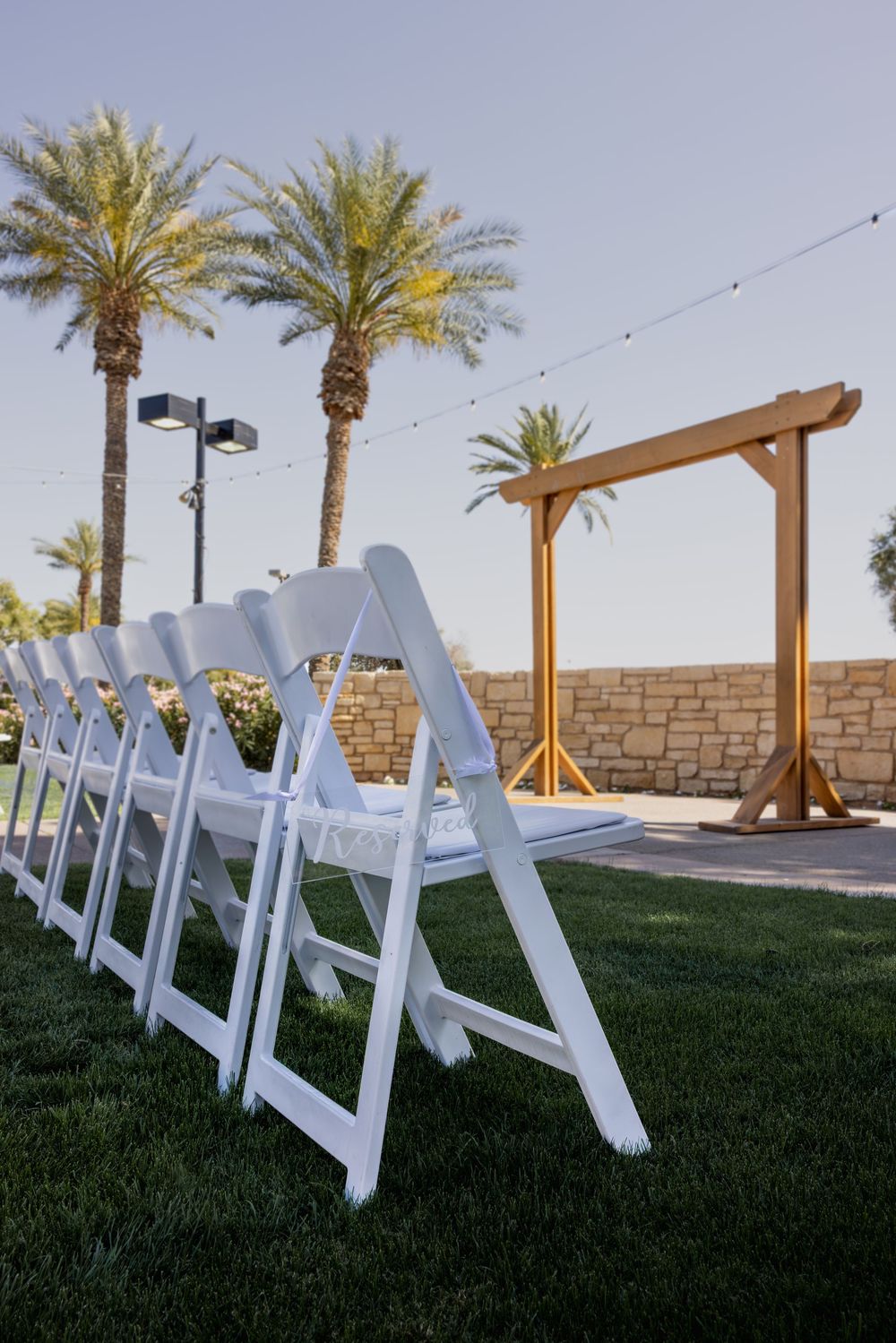 White folding chairs arranged on green grass with a wooden wedding arch and palm trees at an outdoor ceremony venue.