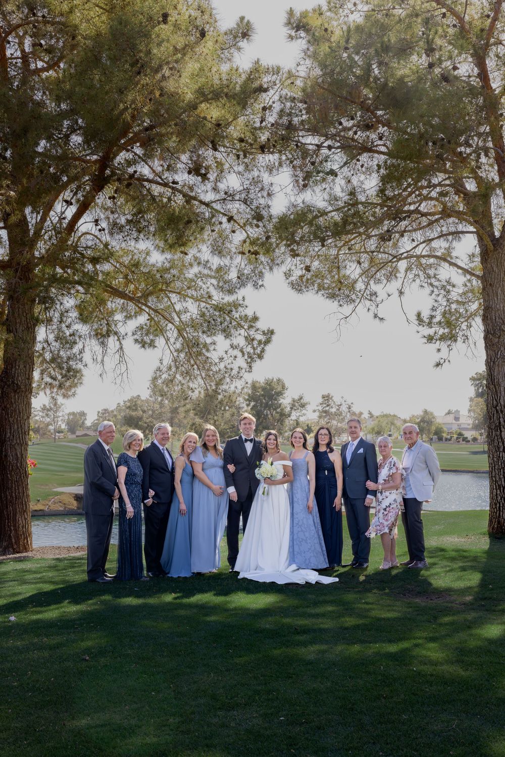 Large wedding family portrait group standing under pine trees beside a pond on a lush golf course.