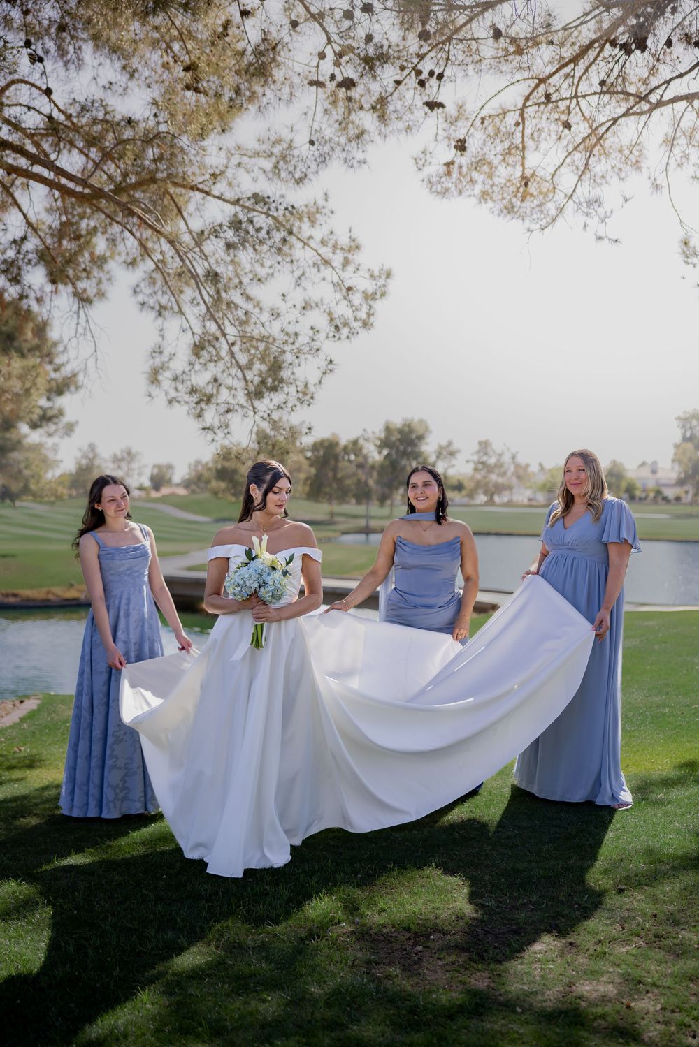 Bride in off-shoulder white gown with three bridesmaids in dusty blue dresses holding bouquets on golf course.