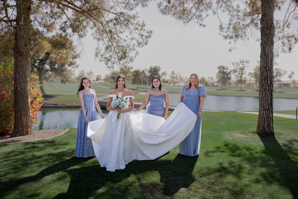Bride and three bridesmaids in blue dresses posing with bouquets on green lawn beside a scenic pond.