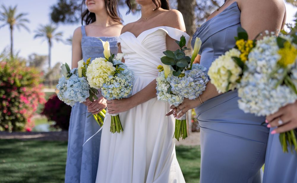 Close-up of bride and bridesmaids holding white hydrangea and blue floral bouquets in dusty blue gowns outdoors.