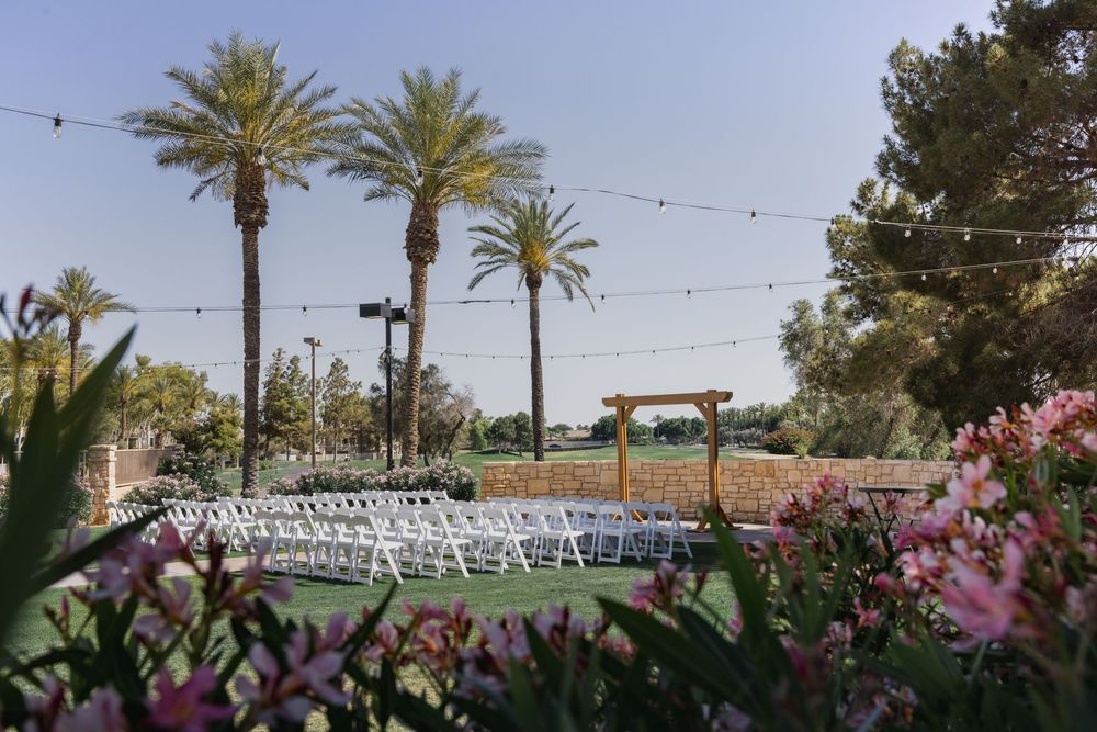 Outdoor wedding ceremony setup with white chairs, string lights, wooden arch, and tall palm trees in warm evening light.