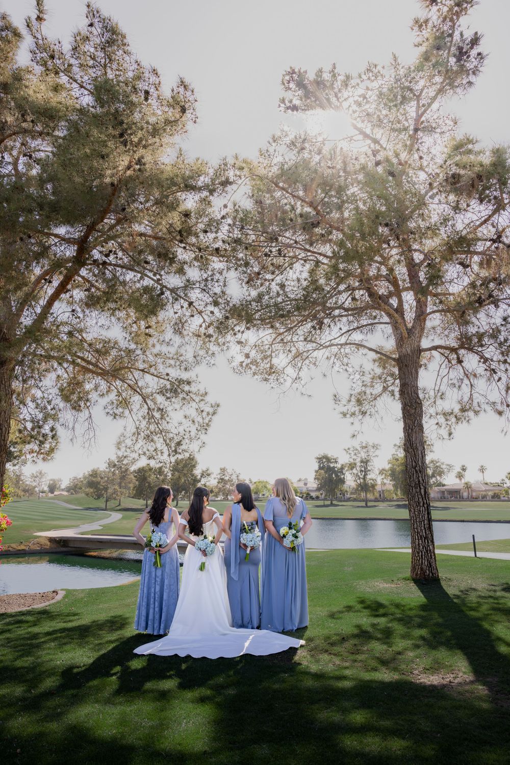 Bride and bridesmaids in blue dresses viewed from behind standing near a pond under tall pine trees.
