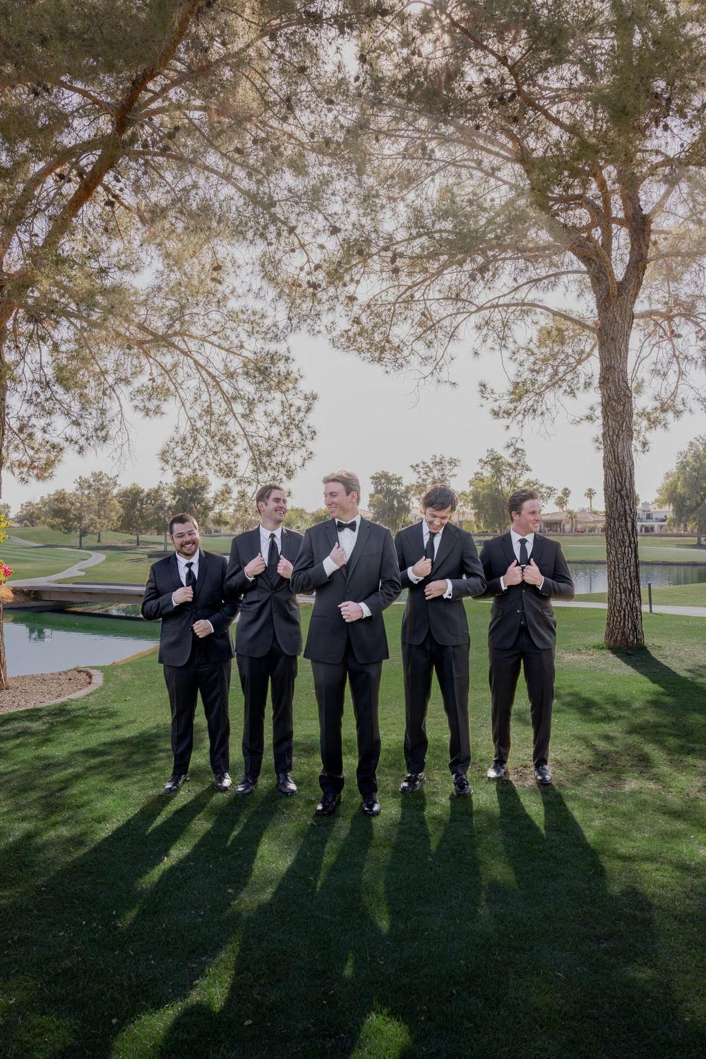 Five groomsmen in black tuxedos adjusting their lapels and smiling on a green golf course lawn.