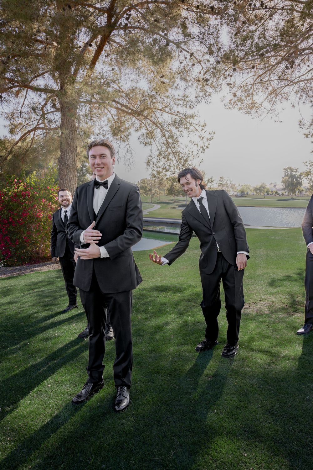 Groomsmen in black tuxedos laughing and walking on green grass at an outdoor wedding venue.