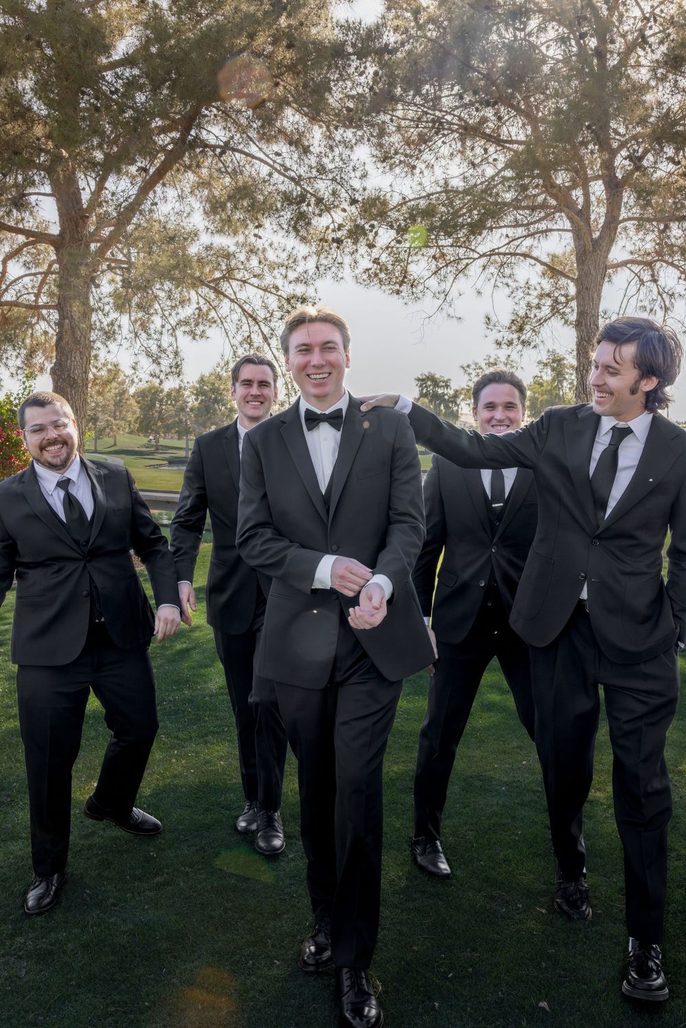 Groom in black tuxedo poses with smiling groomsmen on a green lawn at an outdoor wedding.