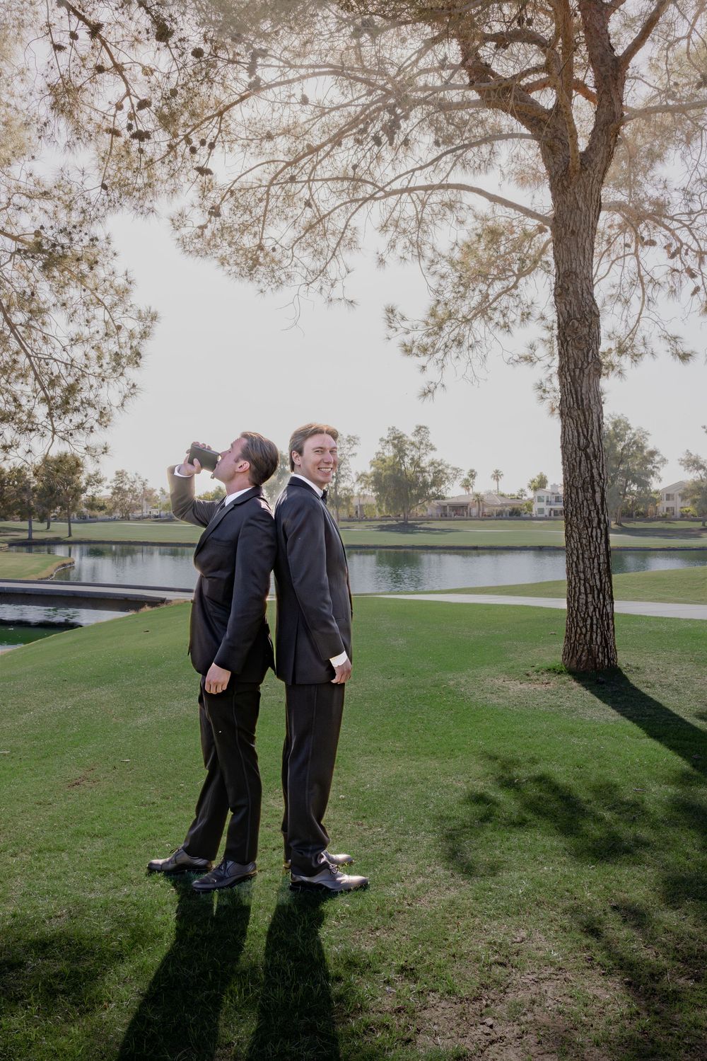 Two men in black suits stand back-to-back near a tree by a pond at a wedding venue.