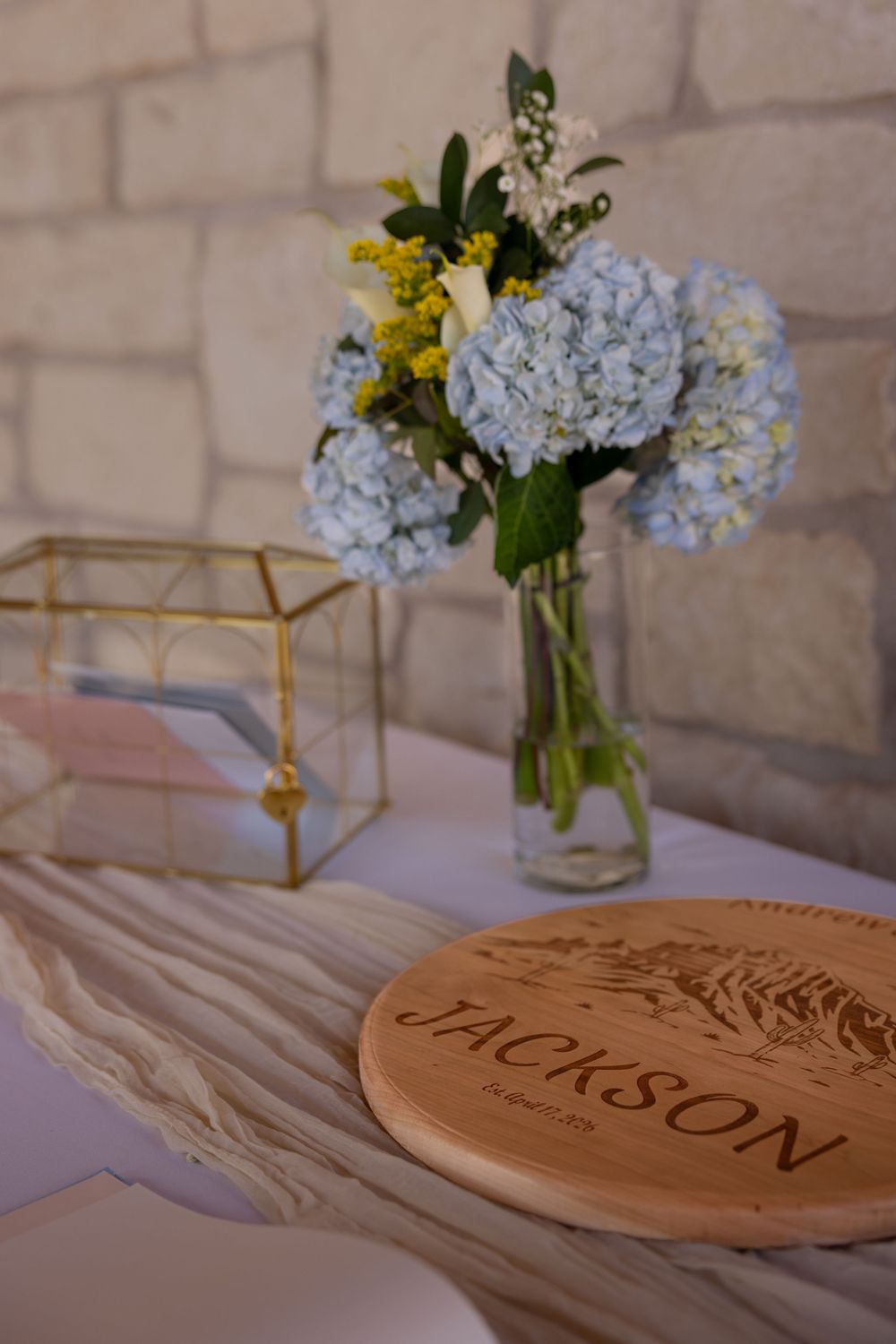 Wedding reception table with blue hydrangea bouquet, gold geometric box, and engraved Jackson wooden sign.