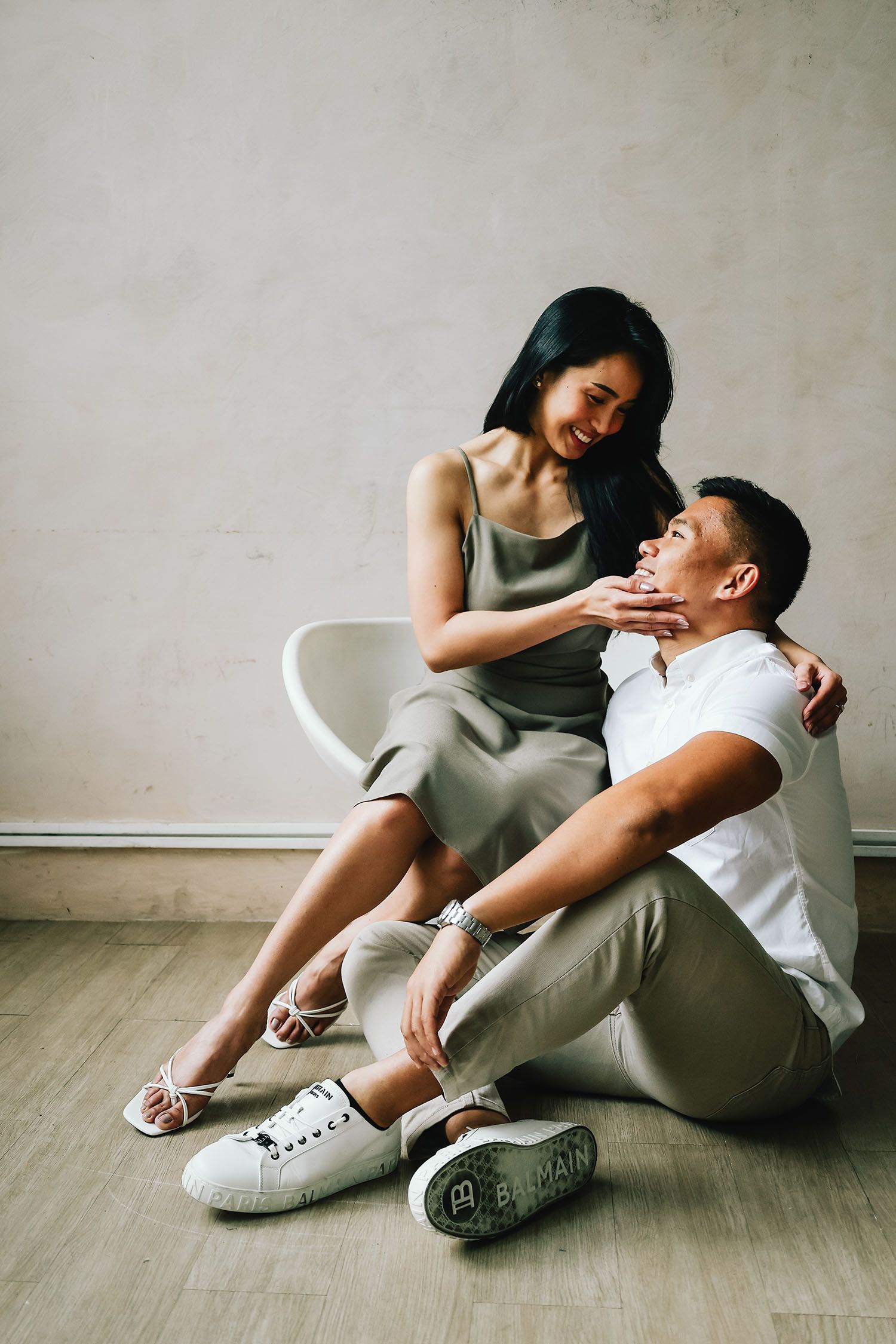A couple in casual attire shares intimate moments while sitting together on a wooden floor in a minimalist setting.