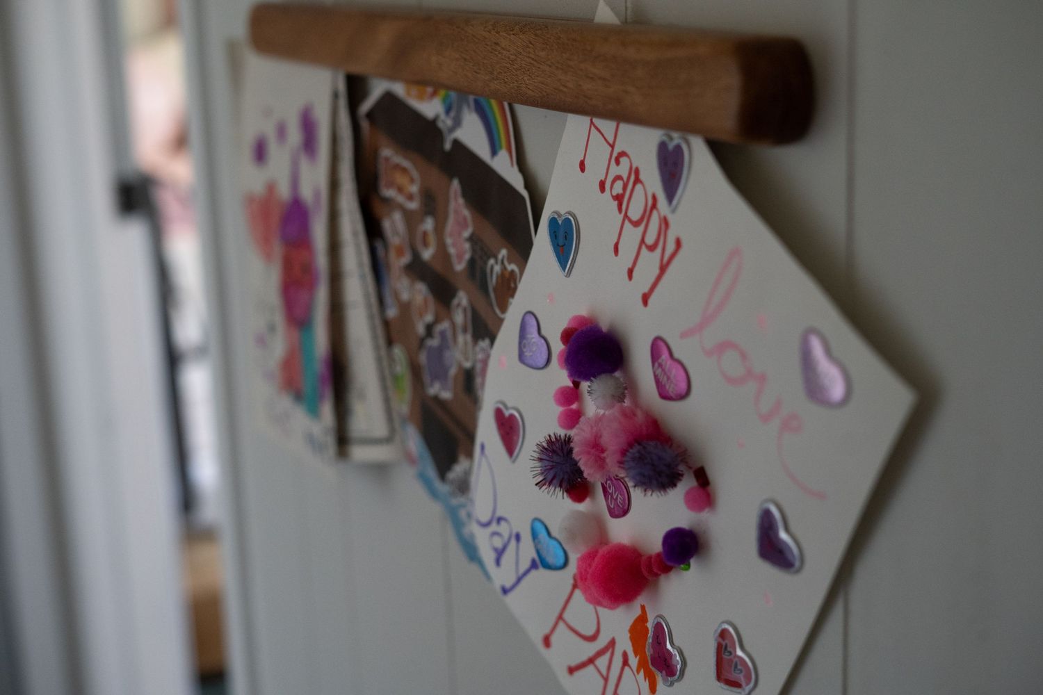 Artwork and photos hang from a wooden hanger against a white wall, with colorful drawings visible in soft focus.