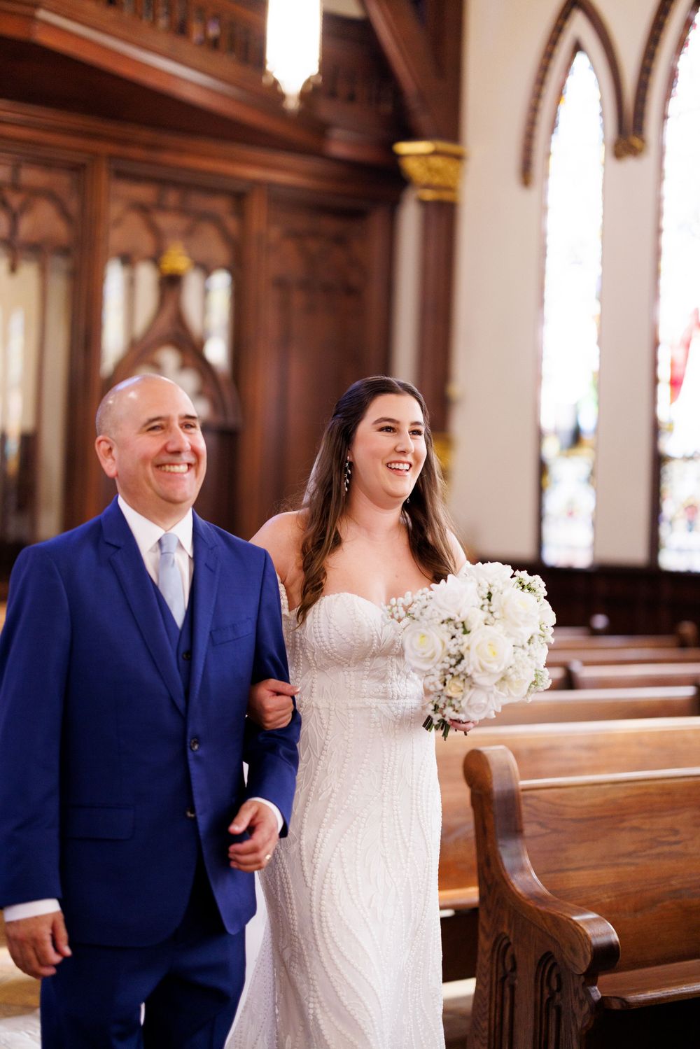 A wedding ceremony processional in a beautiful church with ornate wooden pews and stained glass windows.