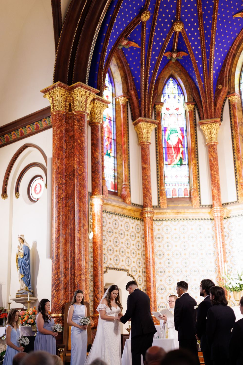 Wedding ceremony in historic church featuring blue painted ceiling, marble columns and stained glass windows.