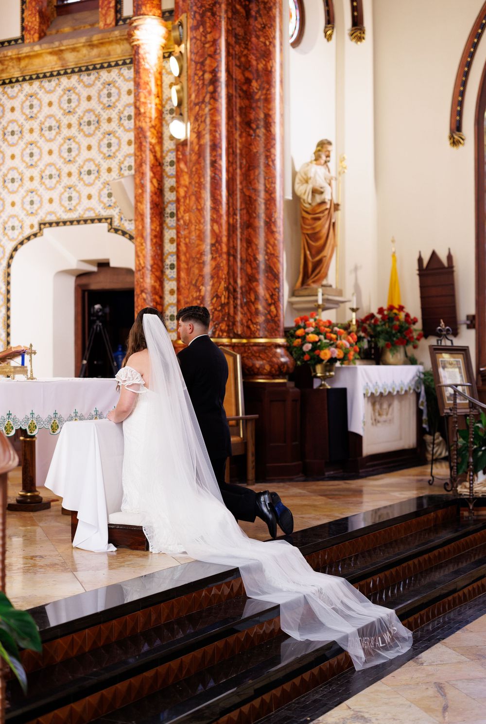 Wedding couple at altar with flowing veil and ornate church architecture in background.