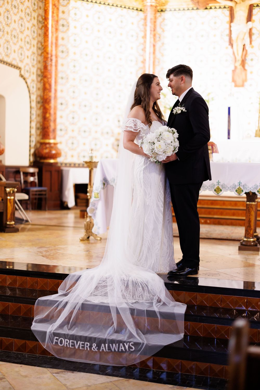 Wedding couple stands on church steps with veil reading 'Forever & Always'.