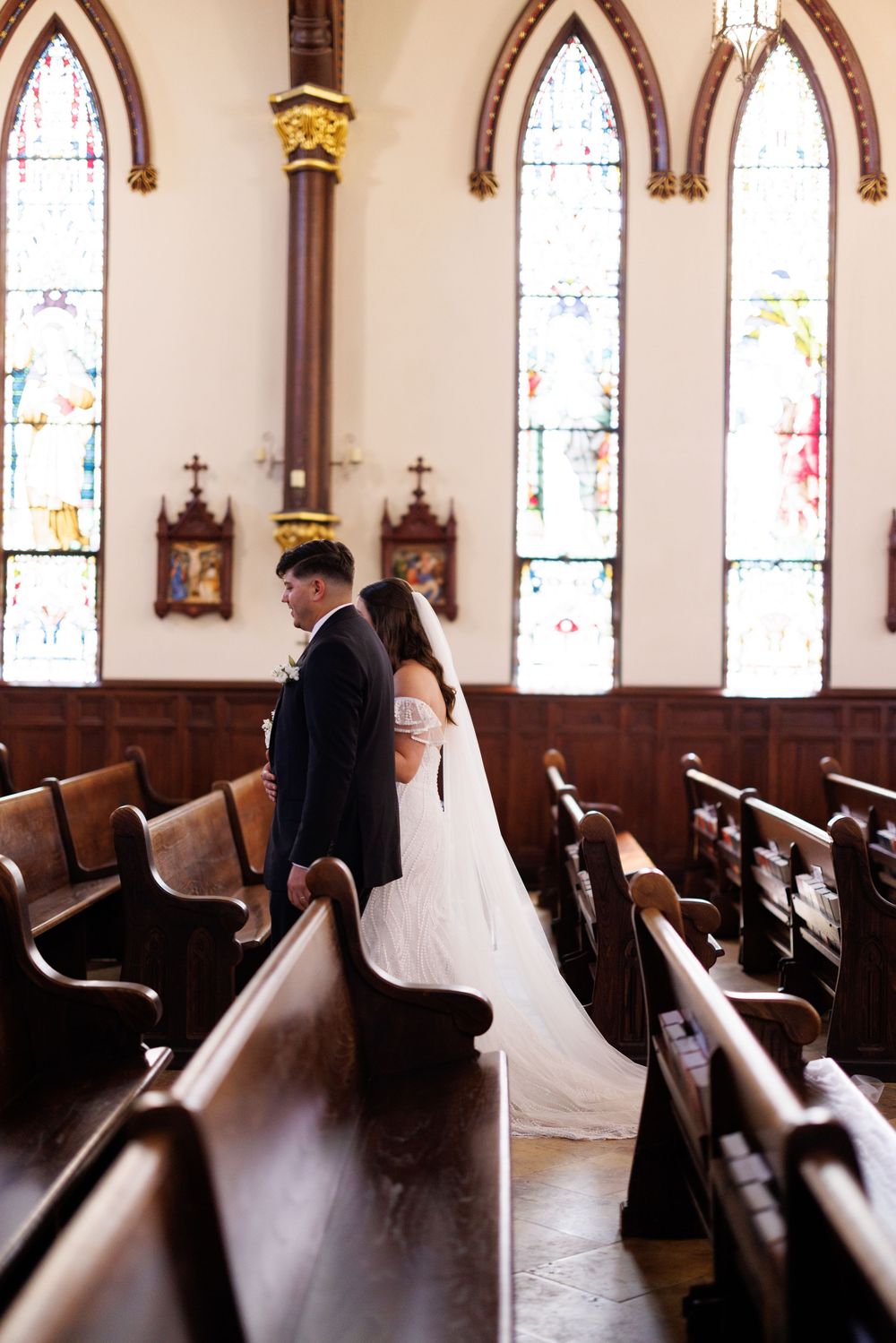 A wedding couple stands in a church aisle with gothic arched stained glass windows and wooden pews.
