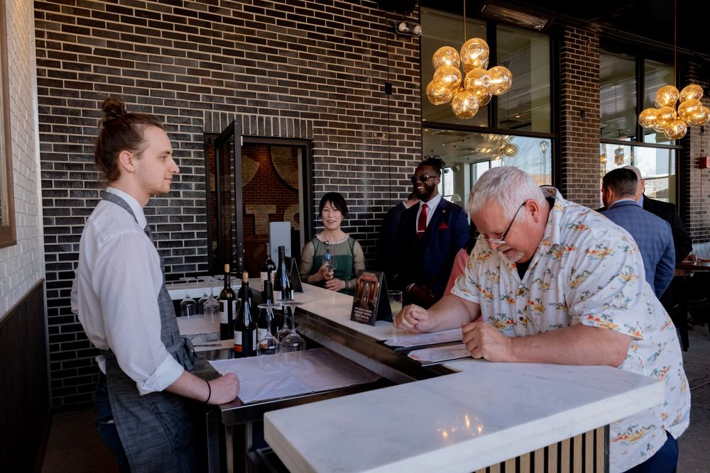 Guests check in at a modern brick-walled restaurant bar while staff assist in a warmly lit dining space.