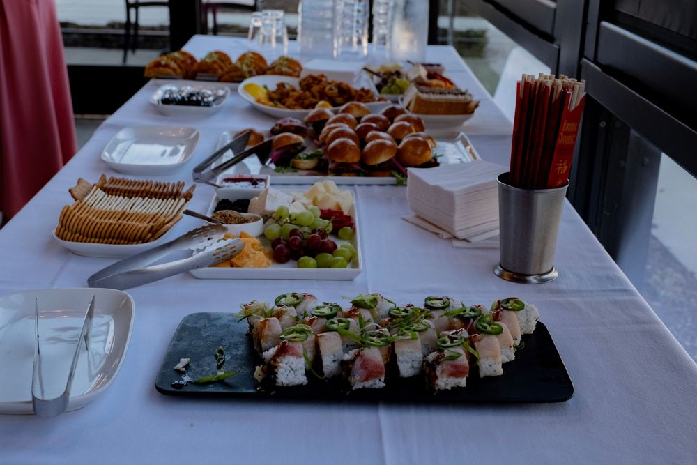 A catered buffet table with sushi rolls, fresh fruit, pastries, and appetizers elegantly displayed on white linen.