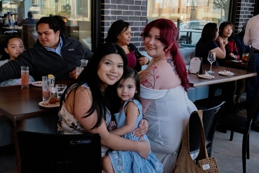 Two women and a young girl pose together smiling at a lively restaurant patio gathering.