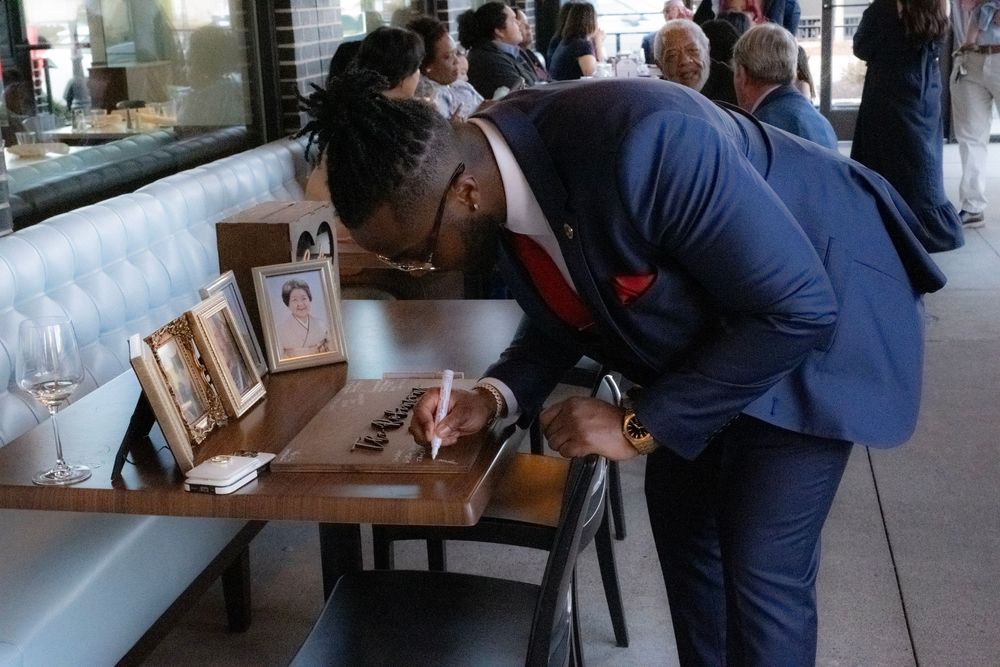 Man in blue suit signing a guest book at a restaurant event, leaning over a table with a framed photo nearby.