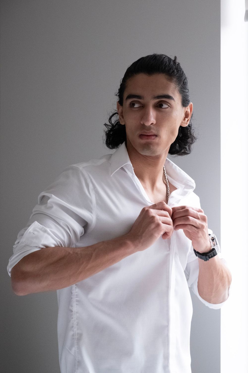 Young man with dark curly hair buttoning white dress shirt while looking sideways near bright window.