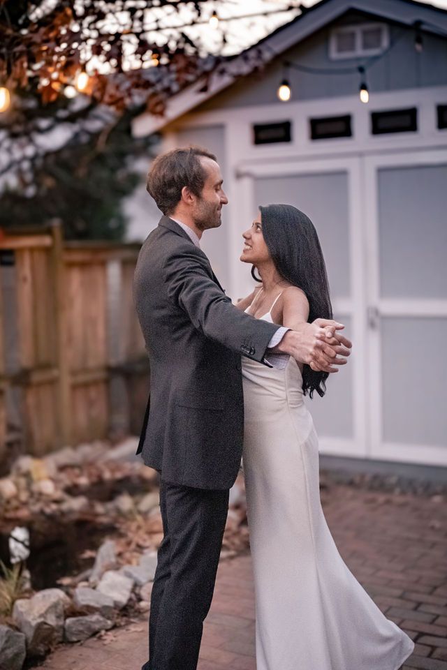 A couple shares a romantic dance outdoors at dusk, the bride in a white gown and groom in a gray suit.