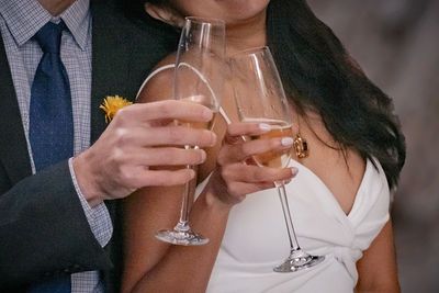 Couple toasting with elegant wine glasses, woman in white dress with gold ring, man in blue suit with yellow boutonniere.