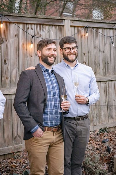 Two smiling men holding champagne glasses at an outdoor backyard gathering with string lights and wooden fence.