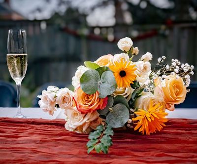 Elegant floral bouquet with orange, peach, and white roses beside a champagne flute on a red tablecloth.